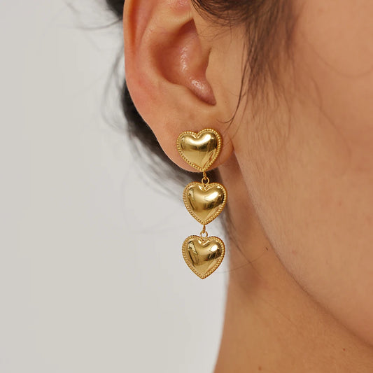A close-up image of a woman's ear wearing a gold-plated heart-shaped drop earring NININOIR 